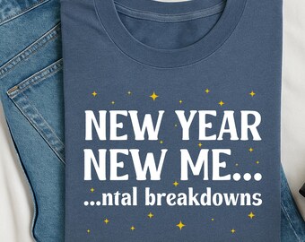 May include: A navy blue t-shirt with the text "NEW YEAR NEW ME... ...ntal breakdowns" in white, with yellow star accents. The shirt is folded on top of blue denim jeans.