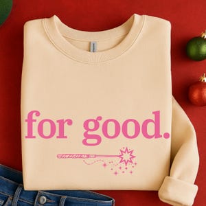 May include: A cream-colored sweatshirt with the phrase "for good." in pink, along with a pink illustration of a magic wand and stars. The sweatshirt is folded, with the sleeves rolled up. A pair of blue jeans is visible at the bottom.