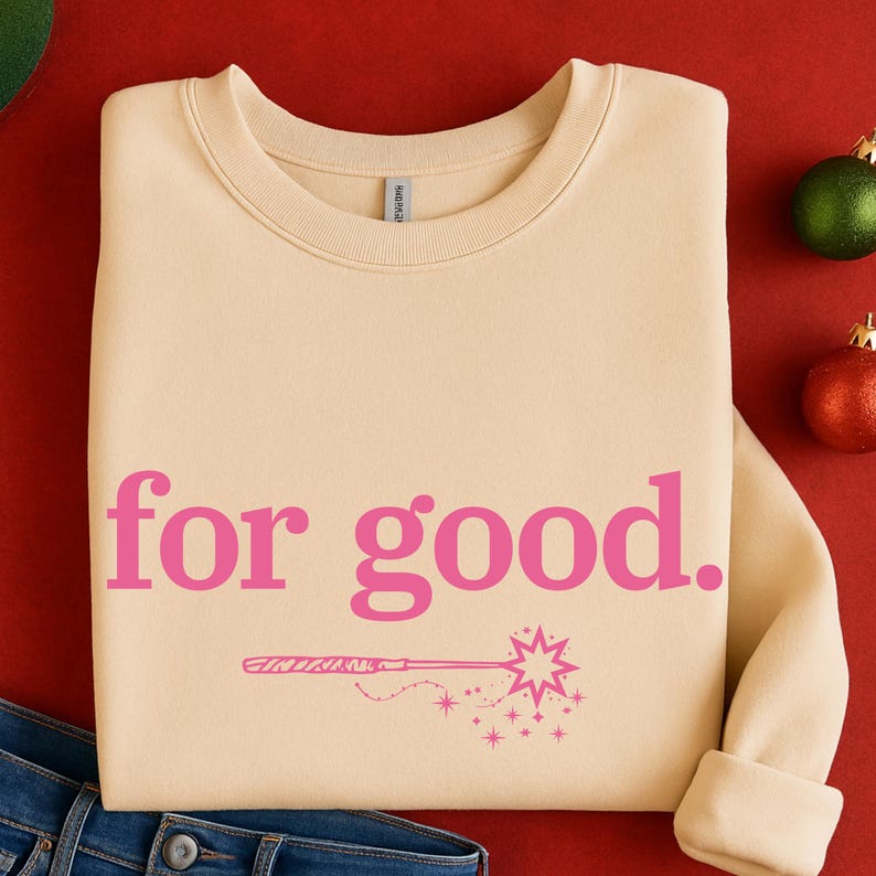 May include: A cream-colored sweatshirt with the phrase "for good." in pink, along with a pink illustration of a magic wand and stars. The sweatshirt is folded, with the sleeves rolled up. A pair of blue jeans is visible at the bottom.