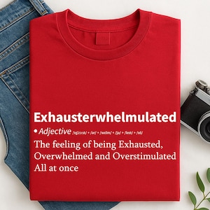 Puede incluir: Una camiseta roja con la palabra "Exhausterwhelmulated" en blanco, con una definición. La definición describe la sensación de estar exhausto, abrumado y sobreestimulado a la vez. La camiseta está doblada sobre unos vaqueros azules.