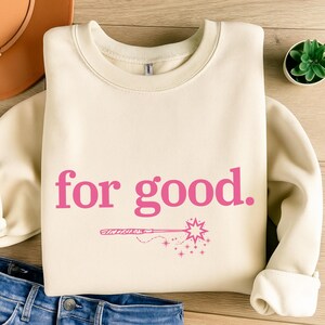 May include: Cream-colored sweatshirt with the phrase "for good" in pink script, accompanied by a pink wand and stars graphic. The long-sleeved top is folded, with a glimpse of blue jeans at the bottom.