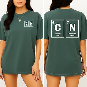 Puede incluir: Camiseta verde oscuro con símbolos blancos de la tabla periódica para carbono y nitrógeno. La parte delantera muestra "C" y "N" en cuadros. La parte trasera muestra los símbolos con números atómicos y pesos, un diseño temático de ciencia.