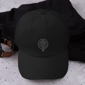 Puede incluir: Gorra de béisbol negra con un gráfico de cerebro blanco. La gorra tiene una visera curva y un diseño clásico de seis paneles. El gráfico del cerebro es una representación estilizada de una placa de circuito.