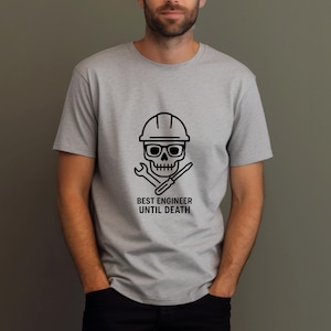 Puede incluir: Camiseta gris claro con un gráfico negro de una calavera con casco y gafas, con una llave inglesa y un destornillador cruzados debajo. El texto "BEST ENGINEER UNTIL DEATH" está impreso debajo del gráfico.