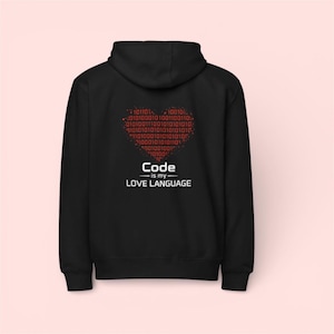 Puede incluir: Sudadera con capucha negra con un diseño de corazón de código binario rojo. El texto "Code is my Love Language" está impreso debajo del corazón. La sudadera tiene capucha y bolsillo delantero.