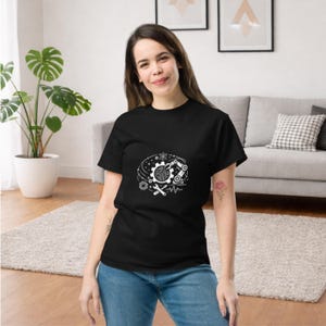 Puede incluir: Camiseta negra con un diseño gráfico blanco que presenta engranajes, una placa de circuito y otros elementos mecánicos. El diseño está centrado en el pecho. La persona lleva vaqueros azules.