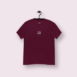 Puede incluir: Camiseta burdeos con un pequeño portátil blanco y un gráfico de símbolo de código en la parte delantera. La camiseta de manga corta cuelga de una percha negra sobre un fondo rosa claro. La camiseta es de color liso.