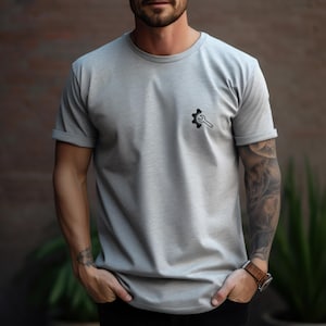 Può includere: Una maglietta grigio chiaro con una piccola grafica nera di ingranaggio e chiave inglese sulla parte superiore sinistra del petto. La maglietta ha maniche corte e girocollo. La persona indossa un orologio in pelle marrone.