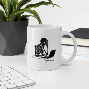 Puede incluir: Taza de cerámica blanca con una ilustración negra de un esqueleto usando una computadora portátil, con el texto "JOHN SMITH" debajo. El esqueleto se representa con la mano en la cabeza, lo que sugiere estrés. La taza está sobre una superficie blanca.