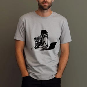 Könnte beinhalten: Hellgraues T-Shirt mit schwarzer Grafik eines Skeletts, das an einem Laptop sitzt und den Kopf in der Hand hält. Das Skelett wirkt gestresst. Das T-Shirt hat kurze Ärmel.