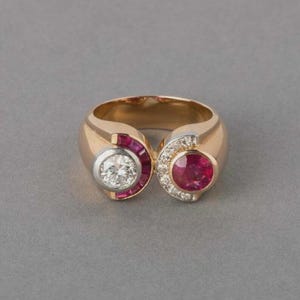 May include: A gold ring with a central diamond and a ruby, each encircled by smaller gems. The ring has a distinctive, open design with a polished finish, set against a grey background.