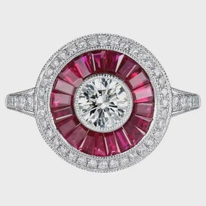May include: A silver ring with a central, round, clear diamond surrounded by a radial pattern of red gemstones. The ring is further embellished with a halo of smaller, clear diamonds, creating a sparkling, elegant design. The band is also adorned with small diamonds.