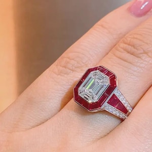 May include: A striking ring with an emerald-cut diamond centre, framed by a red gemstone border. The band is embellished with smaller diamonds, offering a luxurious and captivating aesthetic. The ring's design is elegant and eye-catching.