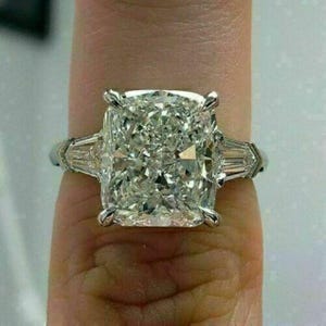 May include: A large, square-cut diamond ring with two smaller, rectangular diamonds on either side. The ring is set in a silver band. The diamond sparkles brightly, reflecting light. A close-up shot highlights the ring's intricate details and brilliance.