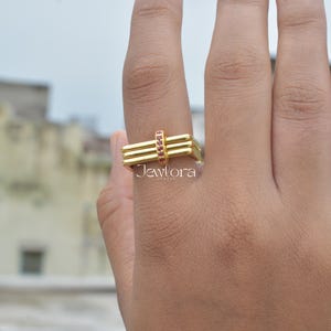 Könnte beinhalten: Ein goldfarbener Ring mit rechteckigem Design. Der Ring hat drei horizontale Balken und einen kleinen Abschnitt mit roten Edelsteinen. Das Wort "Jewlora" ist auf dem Ring aufgedruckt.