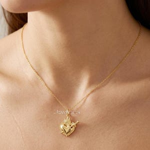 Könnte beinhalten: Eine goldfarbene Halskette mit einem Anhänger in Form eines heiligen Herzens, das von einem Dolch durchbohrt wird und aus dessen Oberseite Flammen lodern. Der Anhänger hängt an einer feinen goldfarbenen Kette. Der Text Jewflora ist sichtbar.