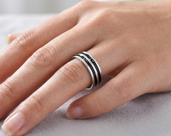 Puede incluir: Un anillo de plata con incrustaciones de esmalte negro. El anillo presenta dos bandas negras separadas por una fina línea plateada. El anillo se muestra en un dedo, destacando su diseño. Una joya sencilla pero elegante.