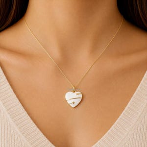 Könnte beinhalten: Eine goldene Halskette mit einem herzförmigen Anhänger, der eine weiße Emaille-Oberfläche und ein goldenes Pfeilmotiv in der Mitte aufweist. Der Anhänger ist mit dem Schriftzug Jewlora in kleinen goldenen Buchstaben oben am Herz versehen.