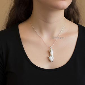 May include: A silver necklace with a pendant shaped like a hand. The pendant is white and silver, with the hand making a fist. The necklace is worn against a black shirt. The brand name "Jewlora" is visible.