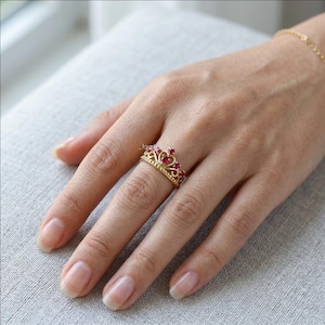 Puede incluir: Un anillo de corona dorado con piedras rojas. El anillo, con forma de corona, presenta detalles de filigrana y múltiples piedras rojas. El anillo está en un dedo.