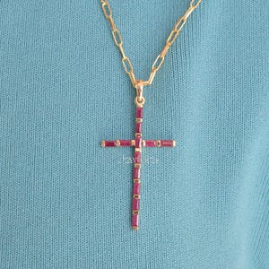 Könnte beinhalten: Eine goldfarbene Kreuz-Anhänger-Halskette mit Kette. Das Kreuz ist mit rechteckigen, tiefroten Edelsteinen besetzt. Die Halskette wird vor einem türkisblauen Hintergrund präsentiert, der das Design und die Farbe des Schmucks hervorhebt.