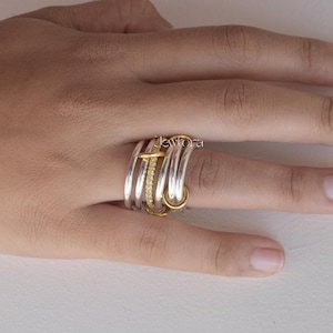 Könnte beinhalten: Eine Ansammlung von Silber- und Goldringen, die auf einem Finger getragen werden. Die Ringe umfassen ein goldenes Band mit kleinen Steinen, einen goldenen Ring mit einem vertikalen Balken und mehrere silberne Bänder. Heller Hintergrund.
