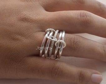 May include: A silver ring with multiple bands and small, sparkling stones. The ring has a unique design with interconnected loops and a polished finish. The ring is on a person's finger.