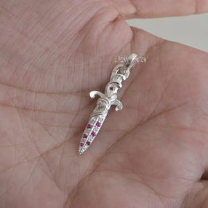 Può includere: Un ciondolo a forma di pugnale d'argento con un manico e una lama decorativi. La lama è adornata con piccole pietre preziose rosse e pietre trasparenti. Il ciondolo è tenuto in una mano. Il testo "Jewlra" è visibile.