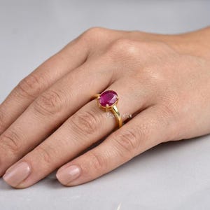 Könnte beinhalten: Ein goldener Ring mit einem großen, oval geschliffenen Rubin. Der Rubin hat eine tiefrote Farbe und ist in einem goldenen Band gefasst. Der Ring wird an einem Finger gezeigt.