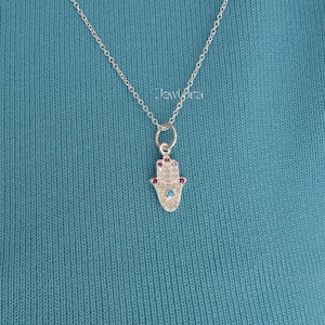 May include: A silver necklace featuring a Hamsa hand pendant. The pendant is adorned with small red and blue gemstones. The necklace is set against a teal background.