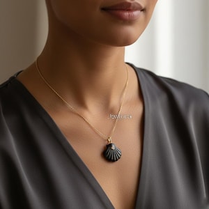 Könnte beinhalten: Eine goldene Halskette mit einem schwarzen Muschel-Anhänger. Der Anhänger hat ein detailliertes Muscheldesign mit einem kleinen, runden, weißen Akzent. Die Halskette wird über einem dunkelgrauen Oberteil getragen.