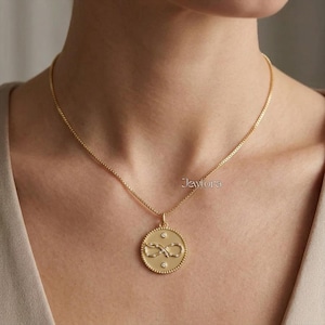 Könnte beinhalten: Eine goldfarbene Halskette mit einem runden Anhänger, der ein Unendlichkeitssymbol und zwei kleine Diamanten zeigt. Der Anhänger hängt an einer filigranen Kette. Die Halskette wird um den Hals getragen.