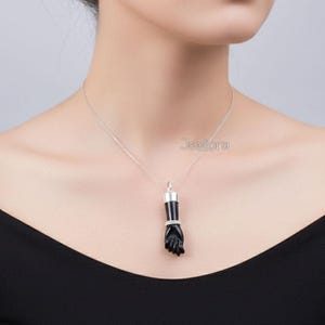 May include: A silver chain necklace featuring a black onyx fist pendant. The pendant has a silver top and a ring of small, clear stones around the wrist. The necklace is worn against a black top.