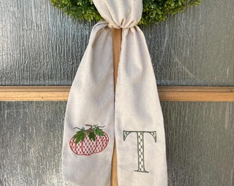 May include: A beige fabric door hanger with a green wreath. The door hanger has a red and green embroidered pumpkin and a green embroidered letter "T". The door hanger is tied to a wooden door frame.