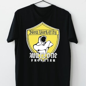 Op de afbeelding: Zwart T-shirt met een gele schildafbeelding. Het schild heeft de tekst "New York City" bovenaan en "Warzone Frontier" onder een witte afbeelding. Het T-shirt hangt aan een zwarte hanger.