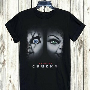 May include: Black t-shirt featuring a graphic of two doll faces with the text "BRIDE OF CHUCKY" in silver lettering. One doll has a blue eye, the other has a green eye. The shirt is hanging on a black hanger.