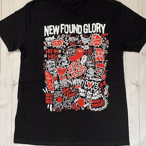 以下が含まれることがあります： 黒いTシャツ。上部に白いブロック文字でバンド名「NEW FOUND GLORY」と書かれています。シャツには、さまざまなフレーズや画像を使った赤と白のグラフィックデザインが施されています。