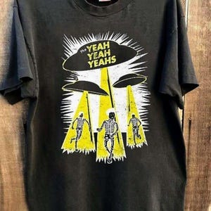 Può includere: T-shirt nera con un design grafico. Il design presenta il testo "The Yeah Yeah Yeahs" sopra tre UFO che proiettano raggi gialli su figure di scheletri. La maglietta è su una gruccia di legno.