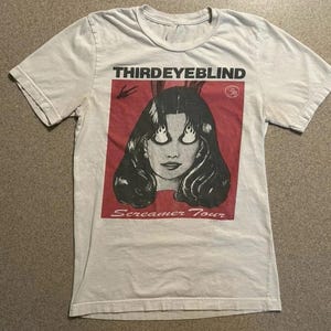 May include: Cream-coloured t-shirt with a graphic of a woman with flame-shaped eyes and the text "THIRD EYE BLIND" and "Screamer Tour" in black and red. The shirt is made of a soft, light-coloured fabric.