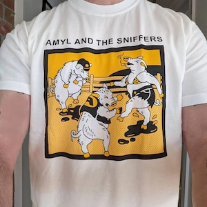 Puede incluir: Camiseta blanca con un gráfico de tres cerdos de dibujos animados en un marco amarillo y negro. Los cerdos están en varias poses, uno de ellos fumando un cigarrillo. El texto "AMYL AND THE SNIFFERS" está encima del gráfico.