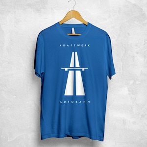 Op de afbeelding: Koningsblauw t-shirt met het woord "KRAFTWERK" boven een witte afbeelding van een snelwegviaduct. Het woord "AUTOBAHN" staat eronder. Het t-shirt hangt aan een houten hanger.