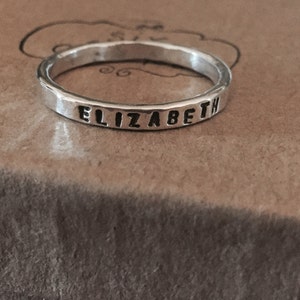 May include: A silver ring with the name "ELIZABETH" stamped around the band. The ring has a hammered texture and sits on a brown surface.