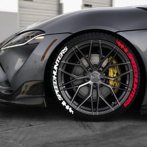 May include: A close-up of a gray sports car wheel with the text "SPEEDHUNTERS" in white and red on the tire. The wheel has a silver rim and a yellow brake caliper. The car is gray.