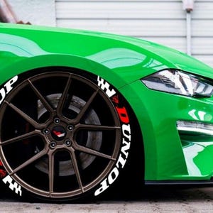 May include: A close-up of a green sports car's front wheel. The bronze-colored wheel has the word "DUNLOP" in white and red lettering on the tire. The car's body is a bright green.