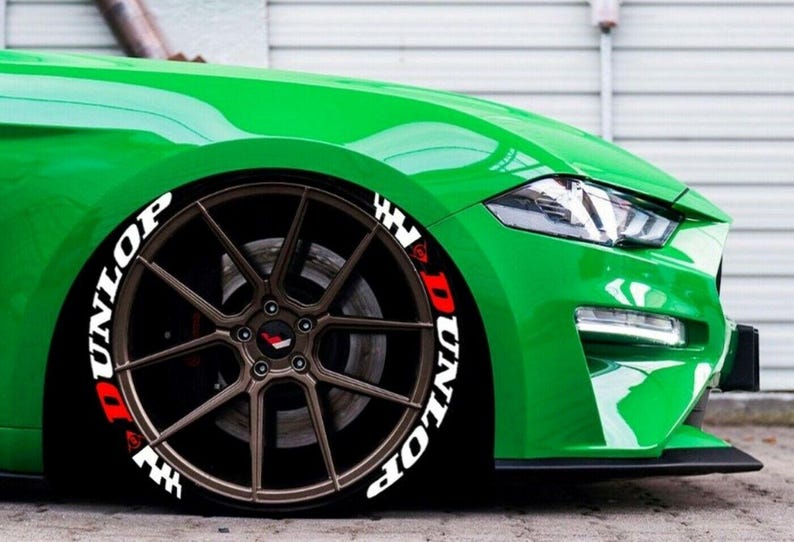 May include: A close-up of a green sports car's front wheel. The bronze-colored wheel has the word "DUNLOP" in white and red lettering on the tire. The car's body is a bright green.