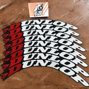 May include: A collection of Dunlop tire stickers. The stickers are black with white text that reads "DUNLOP". The "D" is red. A small sticker with a bear logo and the text "KOM TIRE" is also present.