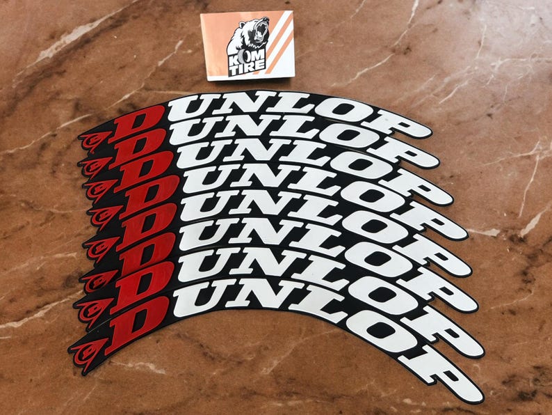 May include: A collection of Dunlop tire stickers. The stickers are black with white text that reads "DUNLOP". The "D" is red. A small sticker with a bear logo and the text "KOM TIRE" is also present.