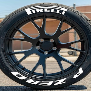 May include: A black car wheel with a tire featuring the white text "PIRELLI" and "P ZERO". The wheel has a multi-spoke design and is set against a blurred outdoor background.