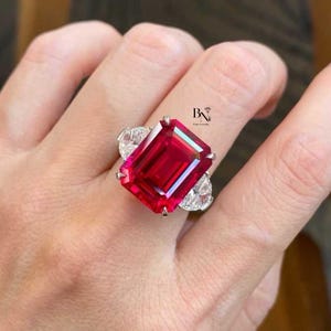 Op de afbeelding: Een opvallende ring met een grote, rechthoekige, levendige rode edelsteen als middelpunt. Twee peervormige, heldere edelstenen flankeren de centrale steen. De ring is gezet in een zilverkleurig metaal.