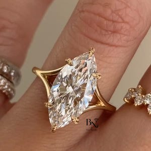 May include: A marquise-cut diamond engagement ring with a gold band. The diamond is large and clear, set in a four-prong setting. The band has a unique, open design. The ring is on a finger.